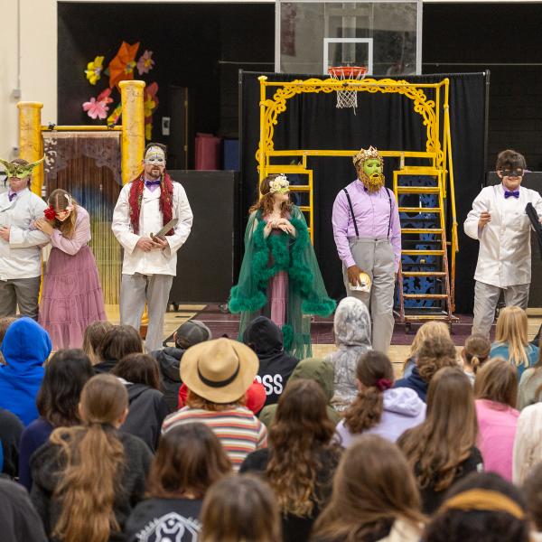 Six actors in elaborate colorful masks perform a Shakespeare play in a school gym 