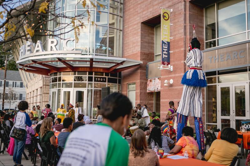 A crowd outside a glass-fronted building, and a person on stilts walking through the crowd.