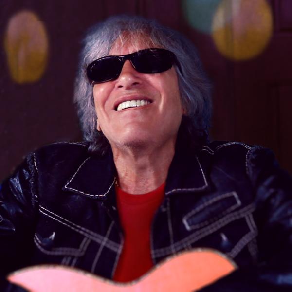 Man with gray hair and dark glasses in blue suitcoat playing guitar.
